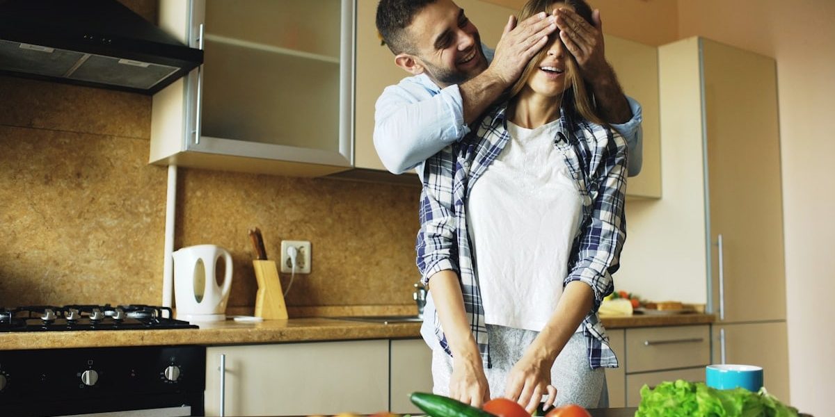 Young man covering girlfriends eyes with hands and surprising her in the kitchen at home. Loving couple cooking breakfast in the morning