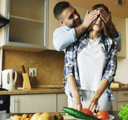 Young man covering girlfriends eyes with hands and surprising her in the kitchen at home. Loving couple cooking breakfast in the morning