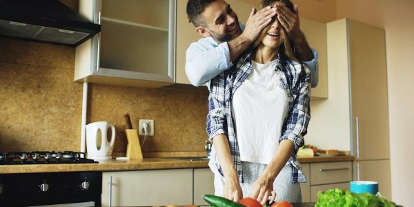 Young man covering girlfriends eyes with hands and surprising her in the kitchen at home. Loving couple cooking breakfast in the morning